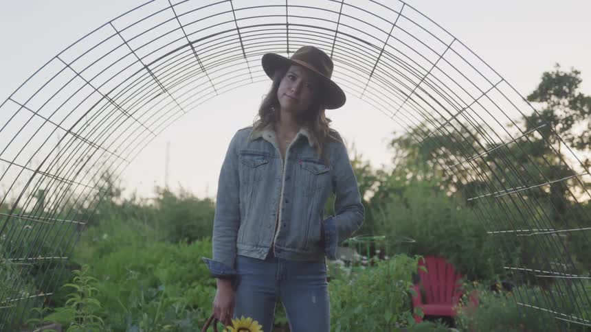 A woman standing outdoors in front of a wire trellis in a garden slowly turns her head to smile into the camera