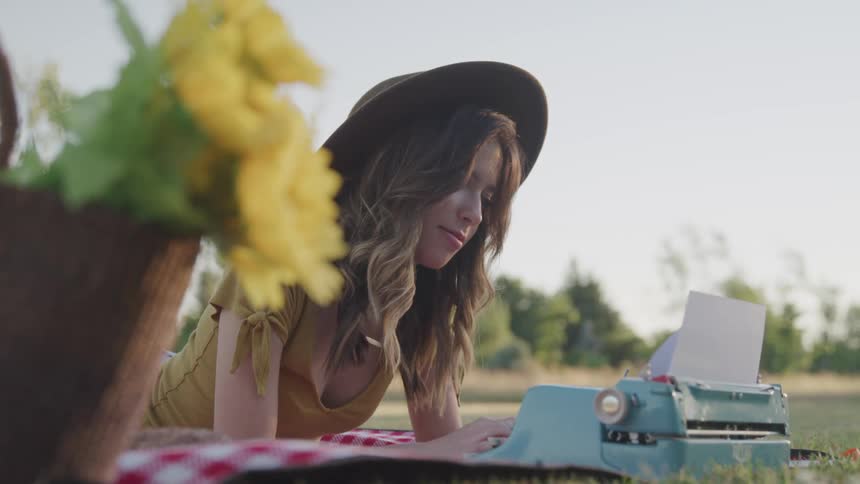 A female lays on a blanket in a field typing on an old typewriter