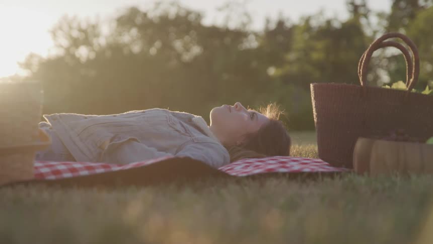 Woman with a denim jacket lies down on a picnic blanket next to the picnic basket at sunset