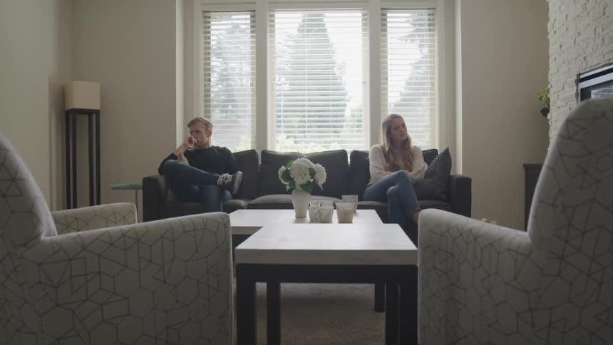Couple sit at opposite ends of couch avoiding looking at each other
