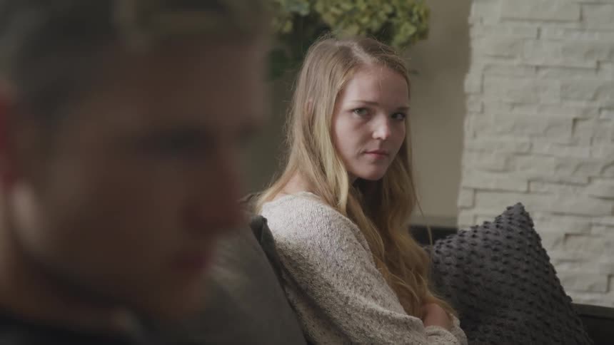 A teenage boy sits on a couch with his head lowered and a teenage girl turns her head and gives him a dirty look