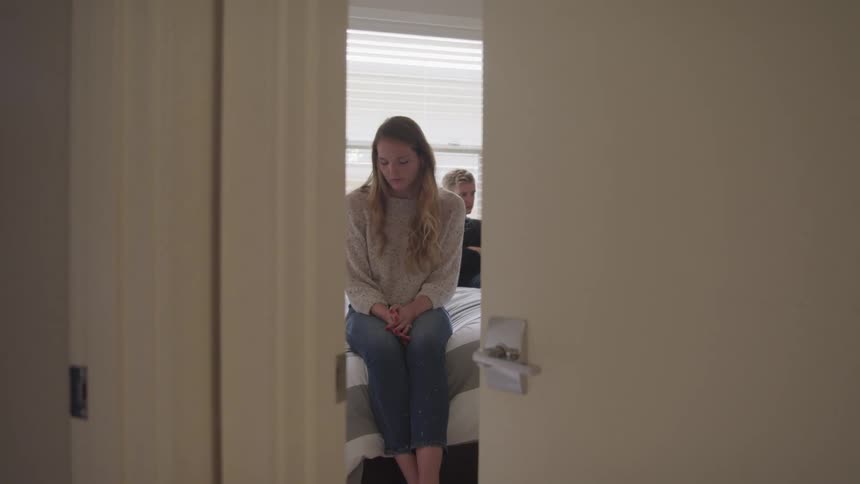A young woman sits on a bed looking glim with a man sitting in a near by chair also looking glim