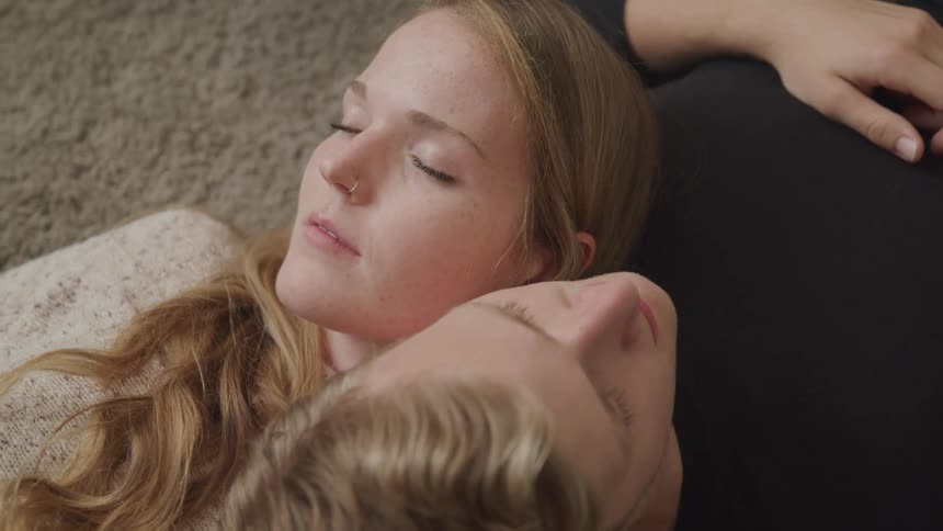 A blonde woman and blond man sleep on carpet facing opposite directions and with their heads resting on each others shoulders