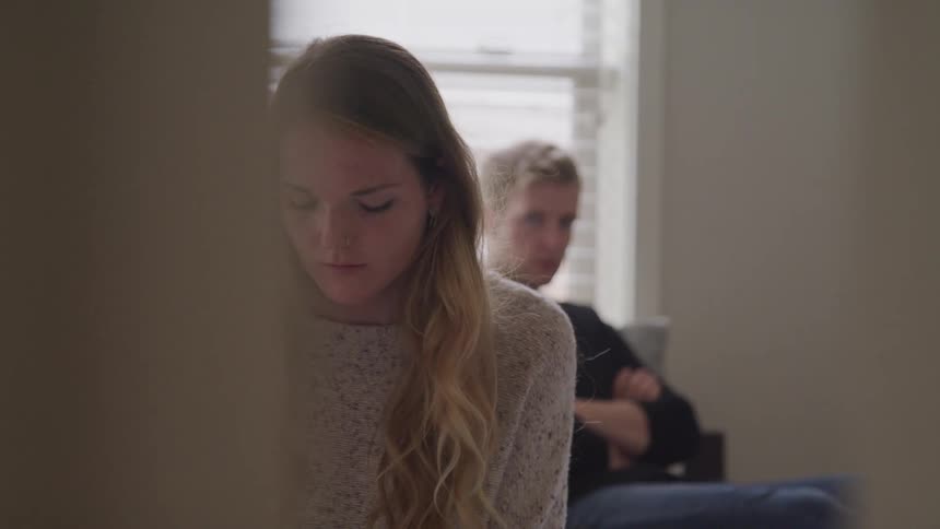 A teenage boy with a scowl on his face sits with his arms crossed and looks at a girl sitting with her back to him