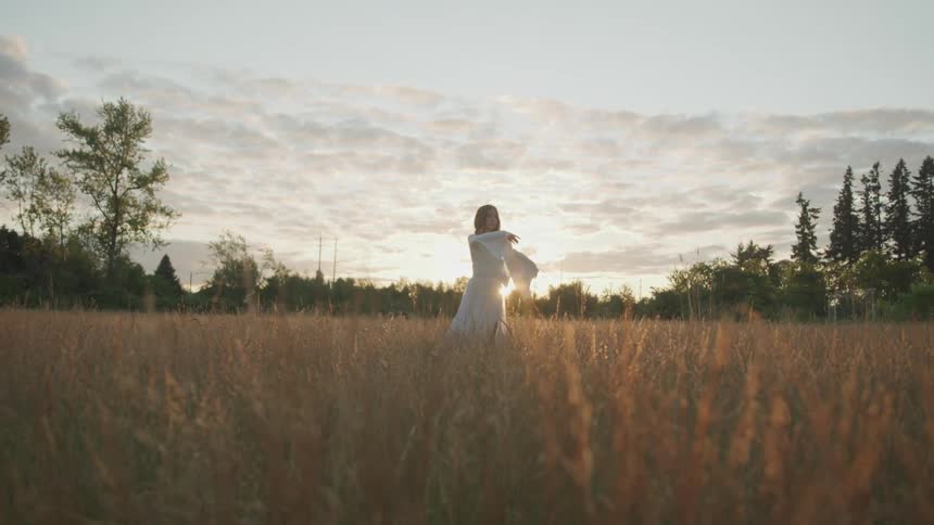 Woman with a white dress dances in tall dry grass in the field at sunset