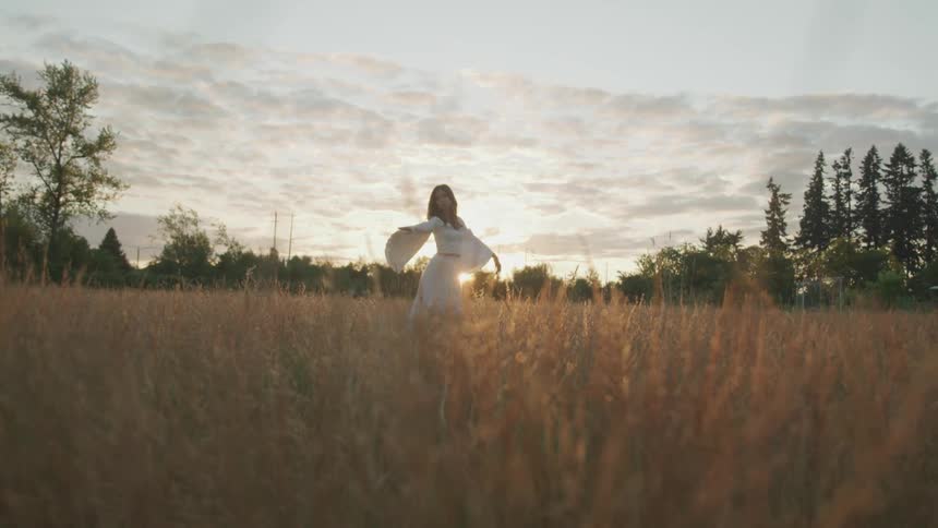 A woman dances in a field of brown grass as the sun peaks over the horizon behind her