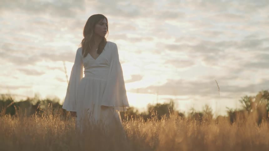 A woman in white clothing stands dramatically in a field of yellow grass as the low sun shines behind her