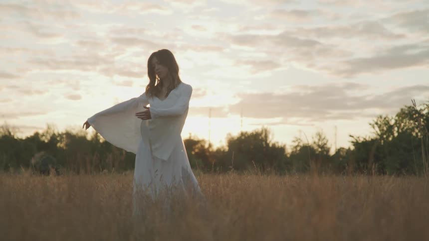 A woman with a white dress dancing in a grassy field with trees and the sky in the background