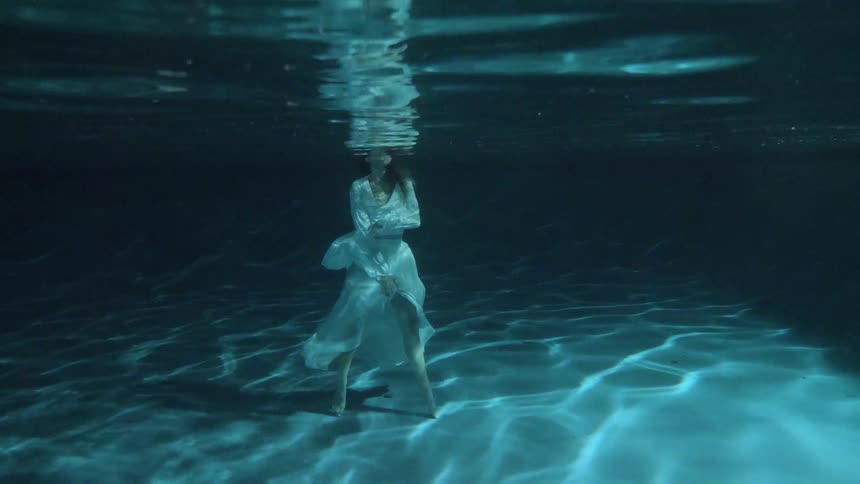 A woman in a white blouse and matching skirt stands in an indoor pool and waves her arms around