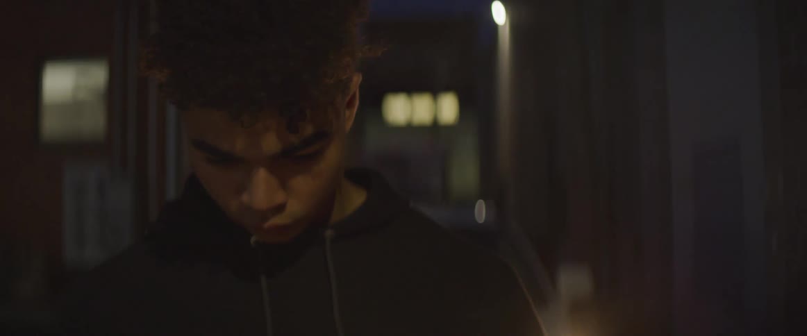 A teenage boy stands in an alley in front of a police car with flashing lights