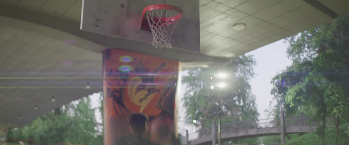 Man plays basketball in covered basketball court in park with foot bridge in background