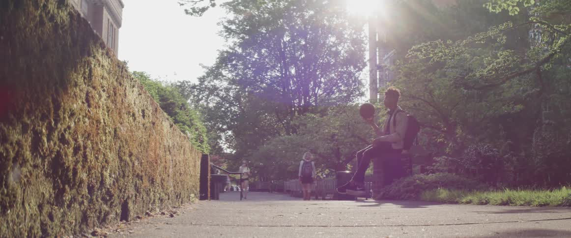 A young man sitting on the side of a walkway outdoors on a sunny day plays with a basketball with his hands as he waits