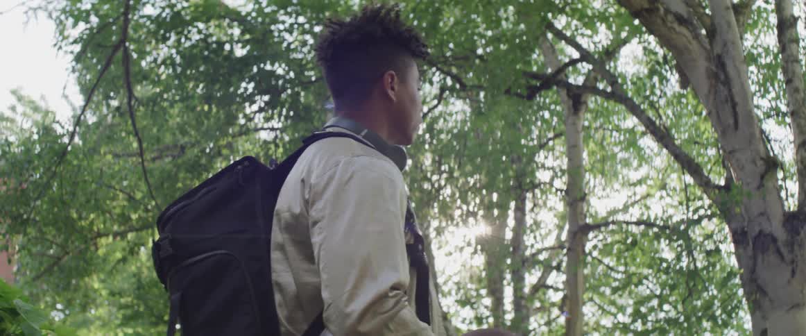 A teenage boys standing under some trees tosses a basketball in the air and catches it