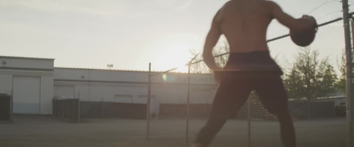 A shirtless black man skillfully dribbles a basketball while making sharp movements outdoors in the evening