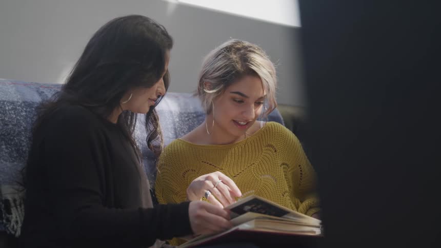 Two women sitting on a couch together look through a book