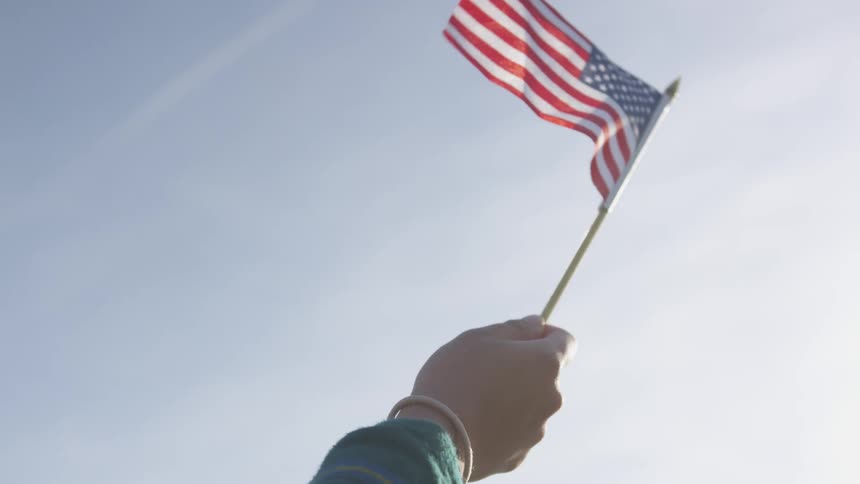 A person holds a small american flag in the wind under a blue sky