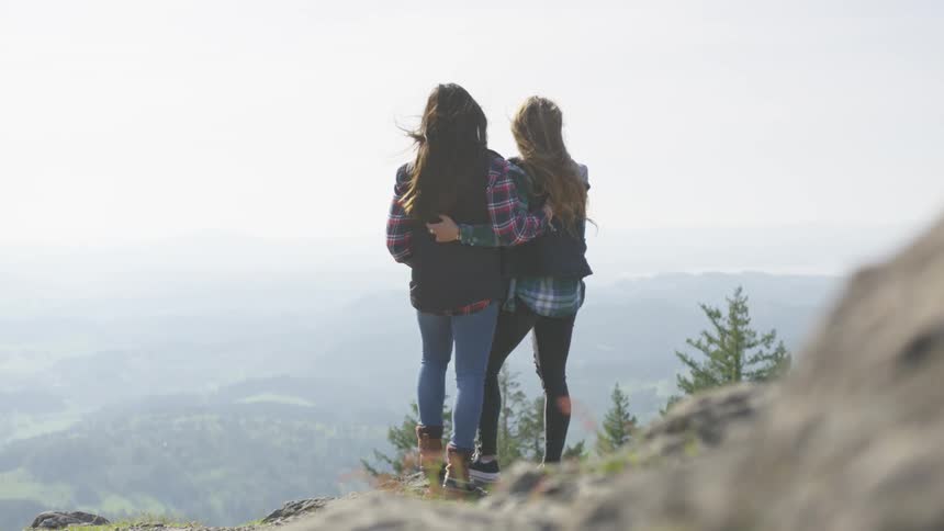Two girls stand on a cliff with their arms around one another and look at the view