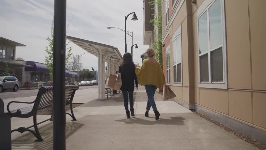 Two friends walk down a city street laughing and carrying shopping bags as a vehicle passes by