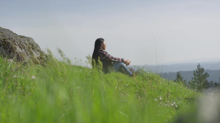 Asian woman with straight hair plaid shirt and jeans sits on the grass on a hill slope