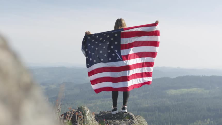 Young woman stands atop a mountain with an american flag extended over her shoulders
