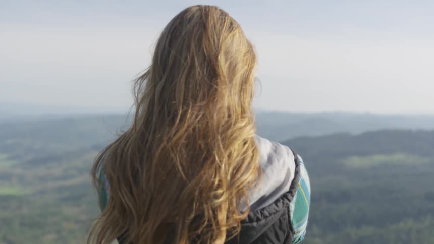 A woman standing at high altitude uses her smartphone to film the forested landscape below