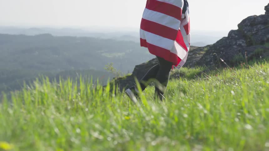 A person draped in an american flag walking up a grassy hill with the sky and clouds in the background