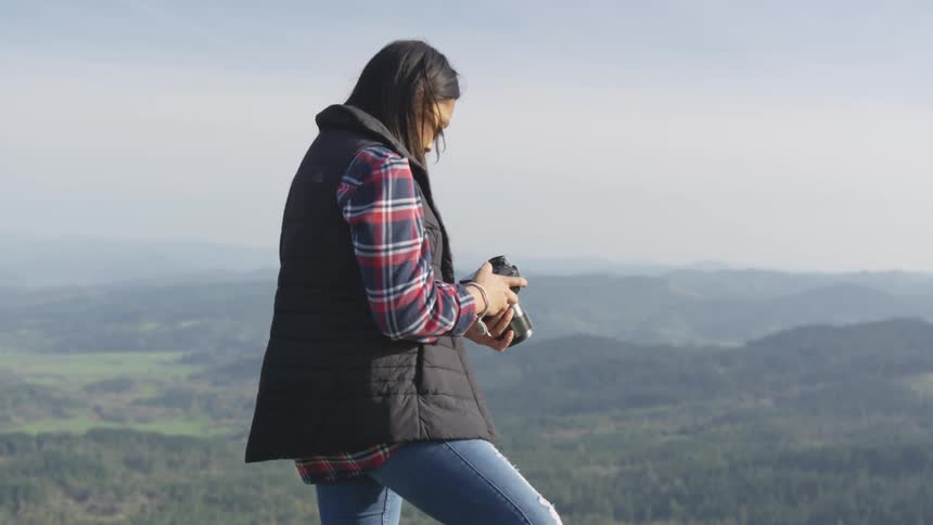 A female photographer standing on a cliff overlooking a countryside covered in forest examines her camera
