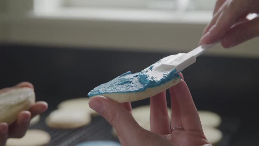 Two people use a spatula to put icing on cookies
