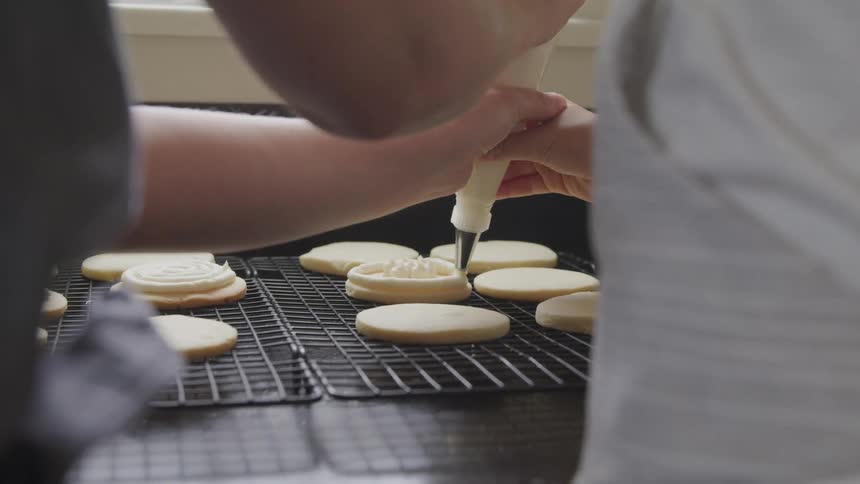 A man and woman work together to squeeze frosting from a pastry bag and decorate a sugar cookie