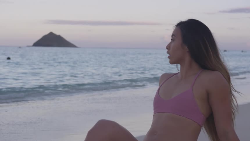Woman wearing bikini reclines on beach as sky on horizon behind islet remains slightly pink