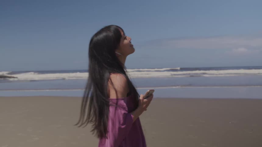 A woman listens to music with earbuds standing on a beach