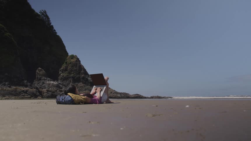 Woman reclines on beach near promontory typing on laptop