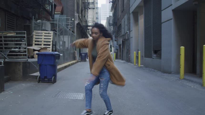 Black woman with curly hair a brown coat and ripped jeans dances in a narrow street