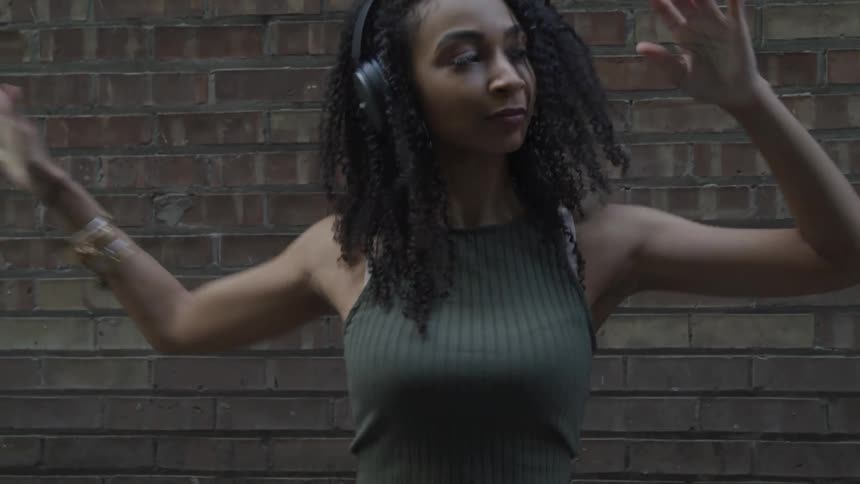 Black woman with curly hair and headphones listens to music and dances in front of a brick wall