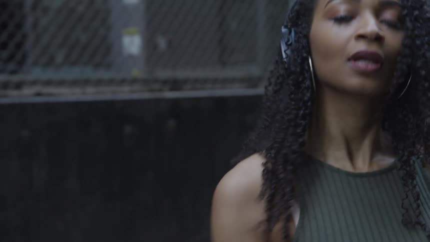 Black female dancer with curly hair hoop earrings and black headphones dances in a narrow street