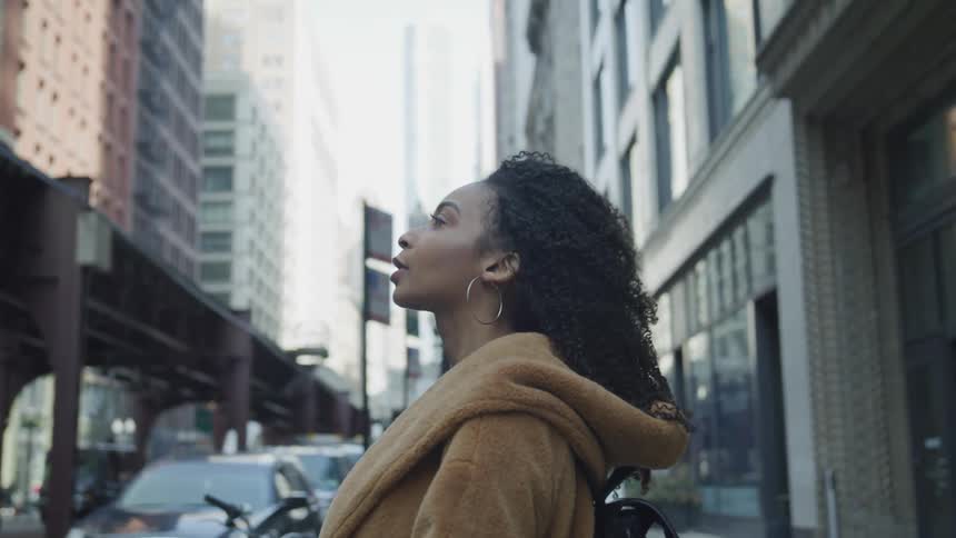 A woman stands on a city sidewalk and looks up to the tall buildings