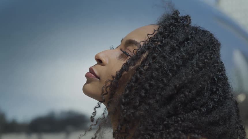 A young black woman stands outdoors facing the daytime sky with her eyes closed