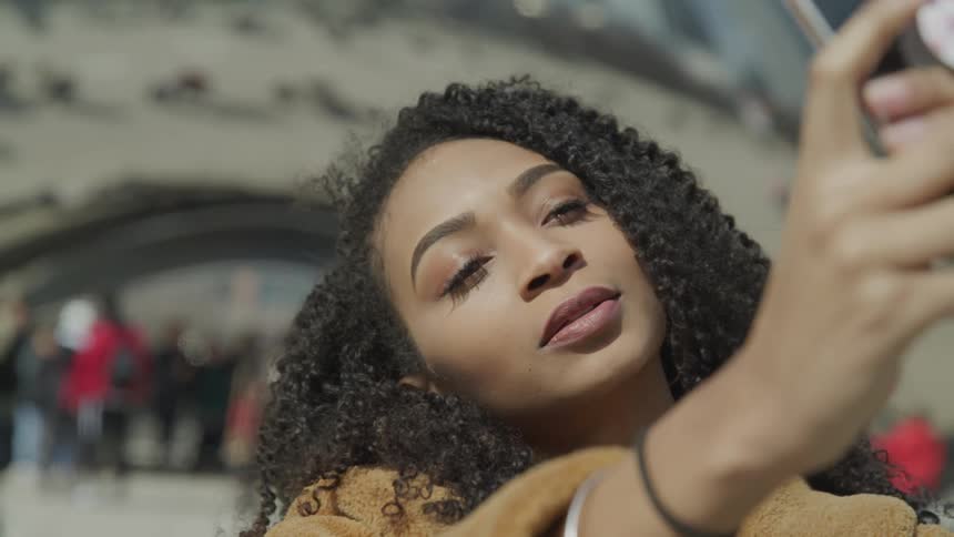 A young black woman poses in front of a landmark as she prepares to take a selfie with her smartphone