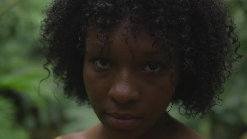 Black woman with a yellow dress and curly hair faces the camera with eye contact with trees in the background