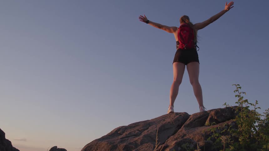 Woman standing on top of large boulder raises arms towards sky