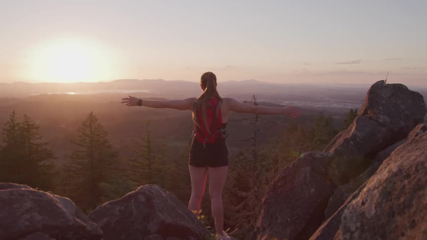 Young woman stands atop a mountain with arms wide open at sunset