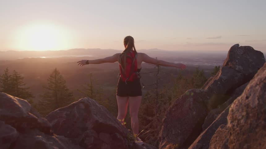 A woman stands on top of a high mountain cliff and spreads her arms in achievement