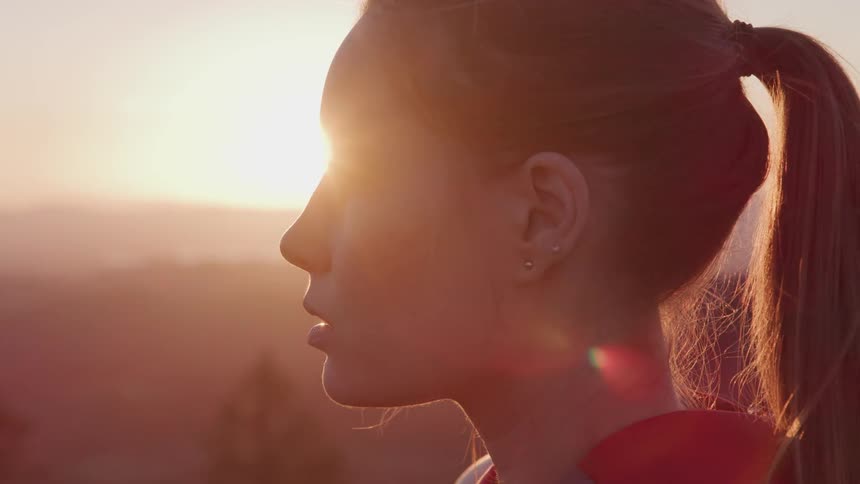 Profile of a young woman standing atop a hill near the pine trees at sunset