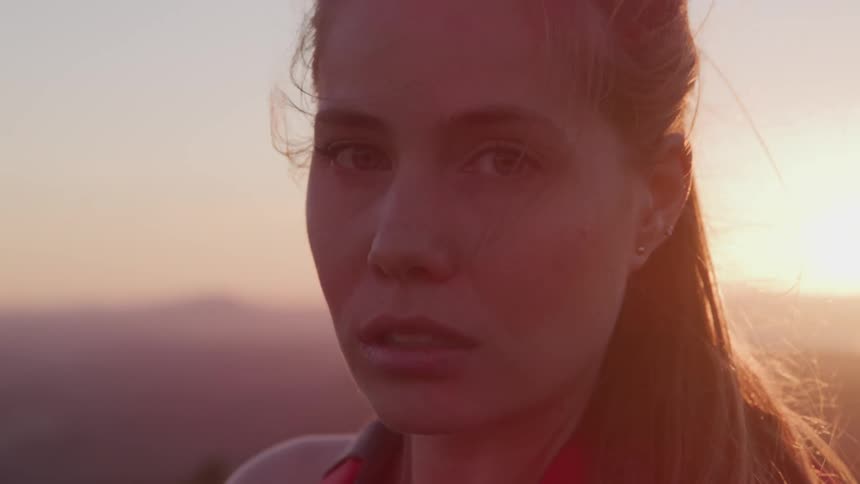 Woman with a ponytail and earrings faces the camera with eye contact at sunset