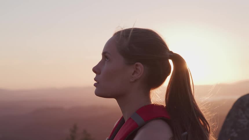 Side view of a woman with a ponytail looks up at the cloudless sky as the sun goes down on the horizon