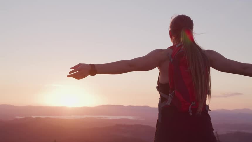 A woman stands atop a mountain with her arms held wide as she faces the setting sun