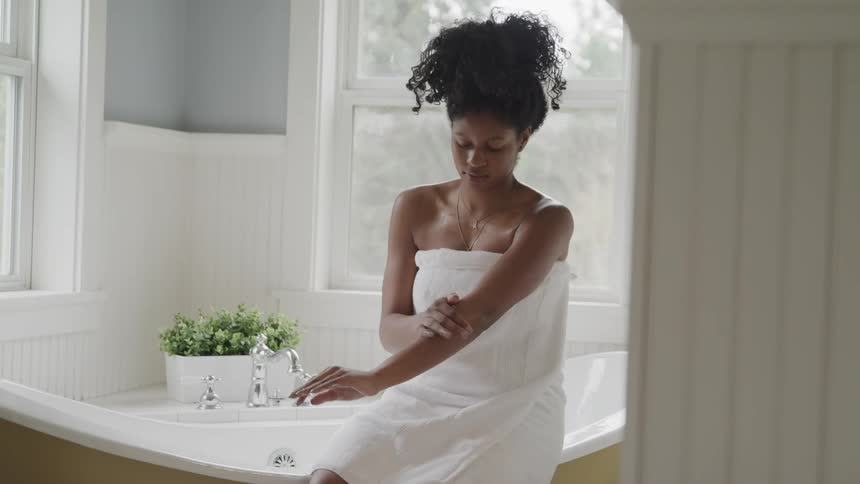 A woman rubs her arm while sitting on the edge of a bathroom wearing only a towel