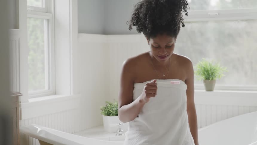 Woman wrapped in towel sits on edge of tub looking at pregnancy test and smiles