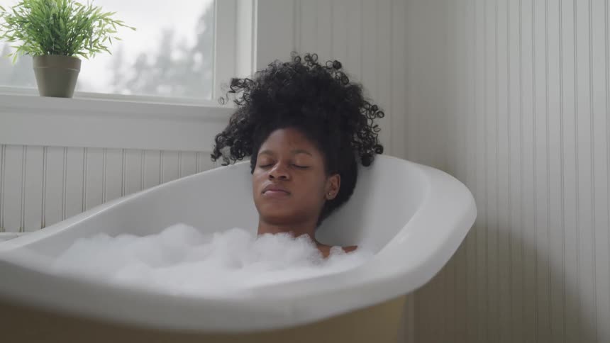 A young black woman rests in a bath tub filled with soap bubbles