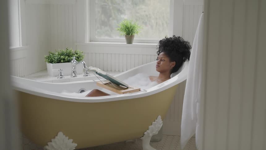 Woman soaks in bathtub reading book propped on holder